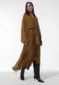 Robe fluide marron à manches longues, avec une taille ceinturée et des accents à franges. Associée à des bottes noires à talons montant jusqu'aux genoux. Texture de tissu lisse.