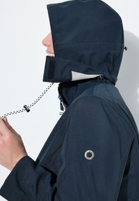 Veste marine imperméable avec capuche, au design minimaliste, dotée d'un cordon de serrage, d'une protection pour le menton et d'un logo circulaire sur la manche gauche.