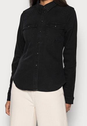 Femme portant une chemise noire ajustée à manches longues avec boutons, poches poitrine à rabat, et un pantalon de couleur claire.