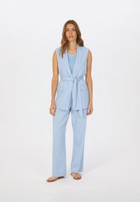 Blazer sans manches bleu clair avec une ceinture à nouer et deux poches avant, assorti à un pantalon ample et à des sandales plates.