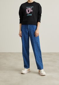 Černá mikina s grafikou "Calvin Klein Jeans" v zelené a růžové; modré široké kalhoty s vzorem; světlé chunky tenisky.