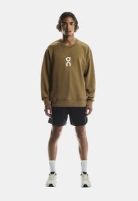 Brun sweatshirt med et hvidt logo foran, kombineret med sorte shorts og hvide sokker. Modellen står foran en hvid baggrund.