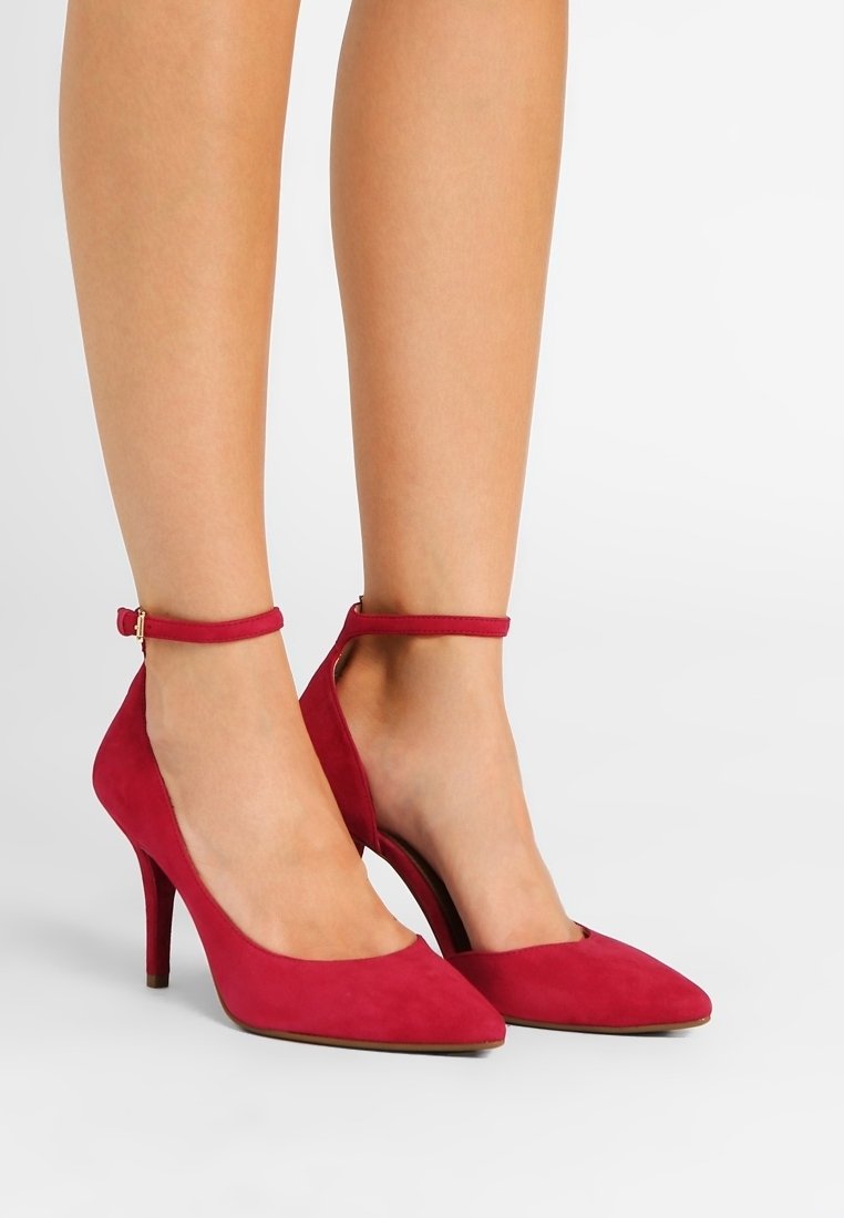 Gambe che indossano tacchi alti a punta in suede rosso con cinturini alla caviglia, ferme su un pavimento bianco.