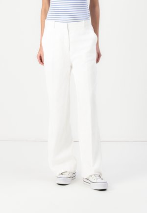 Persona con pantalones blancos de pierna ancha, zapatillas blancas y una camiseta a rayas azul y blanca, de pie sobre un fondo blanco liso.