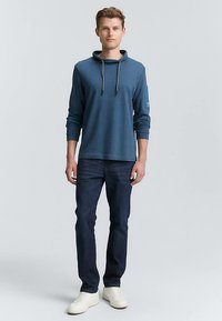 Sweat à capuche bleu avec cordon de serrage, manches longues et une poche sur le bras gauche, porté avec un jean foncé et des baskets blanches.