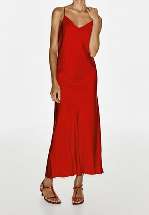 Femme portant une robe longue rouge sans manches à fines bretelles et des sandales rouges à lanières, debout avec une main près de l'épaule.