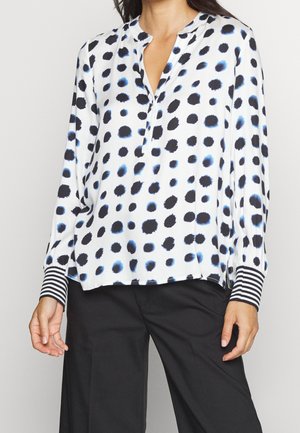 Vrouw draagt een witte blouse met onregelmatige zwarte en blauwe vlekken en gestreepte manchetten, gecombineerd met een zwarte broek.