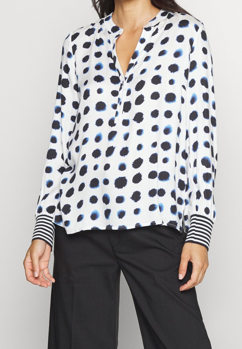 Femme portant un chemisier blanc avec des taches noires et bleues irrégulières et des poignets rayés, associé à un pantalon noir.