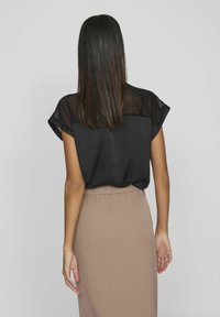 Blusa nera trasparente con maniche corte arrotolate, abbinata a una gonna a tubino beige, caratterizzata da una texture liscia e un design aderente.