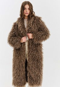 Hnědý kabát z umělé kožešiny s dlouhou, chlupatou texturou, širokými klopami a otevřeným předním dílem. Nošený přes světle zbarvený top a krémové kalhoty.