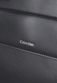 Čierna koženka s viditeľným stehom. Obsahuje strieborné logo "Calvin Klein". Hladký povrch s obdĺžnikovým tvarom a čistými líniami.