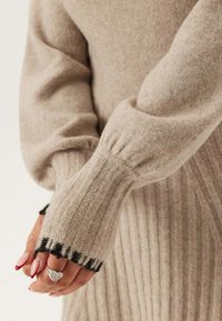 Beige gebreide trui met ribbelstructuur, featuring opgestoken mouwen en zwarte gestreepte manchetten. Een zilveren hart ring op de hand is zichtbaar.