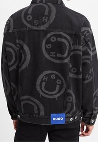 Una persona che indossa una giacca di jeans nera con un motivo circolare grigio e un'etichetta blu "HUGO" sulla schiena, con le mani ai lati.