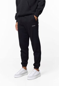 Zwarte joggingbroek van zachte stof, met een elastische tailleband, manchetten en een klein wit logo op de dij. Gecombineerd met witte sneakers.