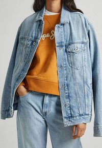 Ljusblå denimjacka med framfickor, silverknappar och sydda detaljer, lager-på-lager över en orange sweatshirt med en vit logotyp.