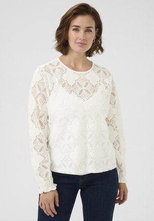 White lace long-sleeve blouse with diamond pattern, round neckline, and ruffled cuffs, paired with dark denim jeans.