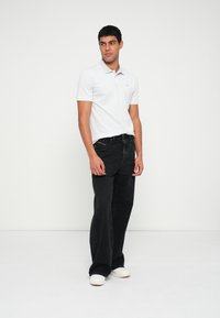 Fehér rövid ujjú galléros póló, fekete bő szárú farmernadrág, és fehér sportcipő. Egyszerű dizájn, finom márkajelzéssel a pólón.