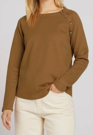 Femme portant un haut marron à manches longues avec quatre boutons sur l'épaule, associé à un pantalon blanc, main dans la poche.