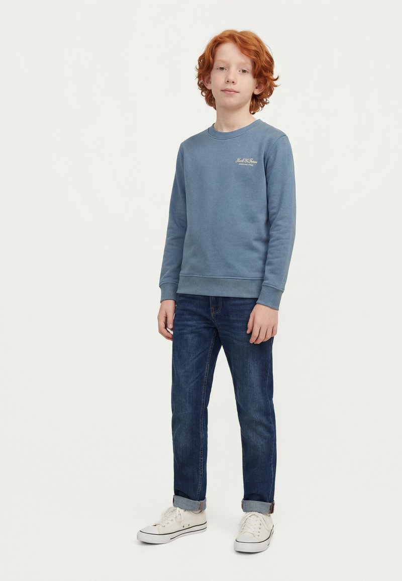 Rødharret dreng stående iført en blå sweatshirt, mørke jeans med opbuk og hvide sneakers mod en enkel lys baggrund.