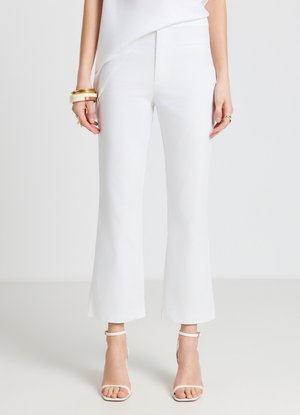 Woman wearing white cropped flare pants with white ankle-strap heels and gold bangles, standing against a plain white background.