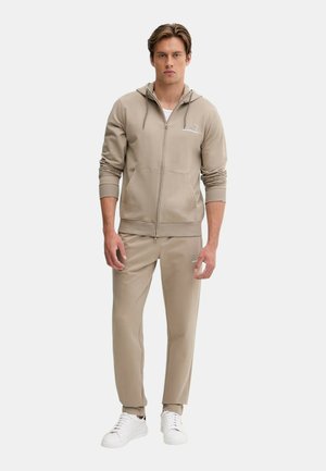 Giovane uomo con felpa beige con zip e pantaloni della tuta abbinati, indossa scarpe da ginnastica bianche, in piedi davanti a uno sfondo uniforme.