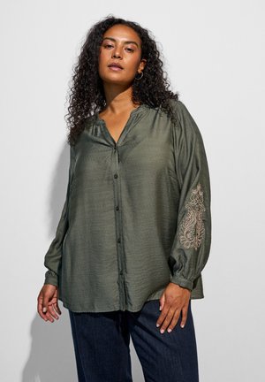 Donna con capelli ricci indossa una camicetta larga verde oliva con bottoni e motivo floreale ricamato sulla manica e pantaloni blu scuro.