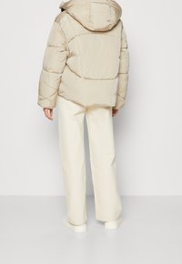 Beige dunjakke med hætte, med sydde sektioner, båret over hvide brede bukser og hvide sneakers. Glat tekstur og afslappet design.