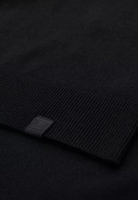 Pull noir avec un corps lisse et un col côtelé, orné d'un subtil patch logo en relief dans le coin. Détail de tissu texturé bien présent.