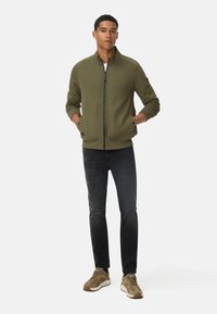 Olijfgroene zip-up jas met gestructureerd ontwerp, ribgebreide manchetten en hoge kraag, gecombineerd met donkere jeans en beige sportschoenen.