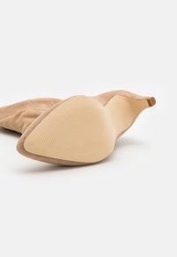 Bottine en daim beige avec un bout pointu et une semelle beige lisse. Présente une texture subtile et un talon bas et arrondi.