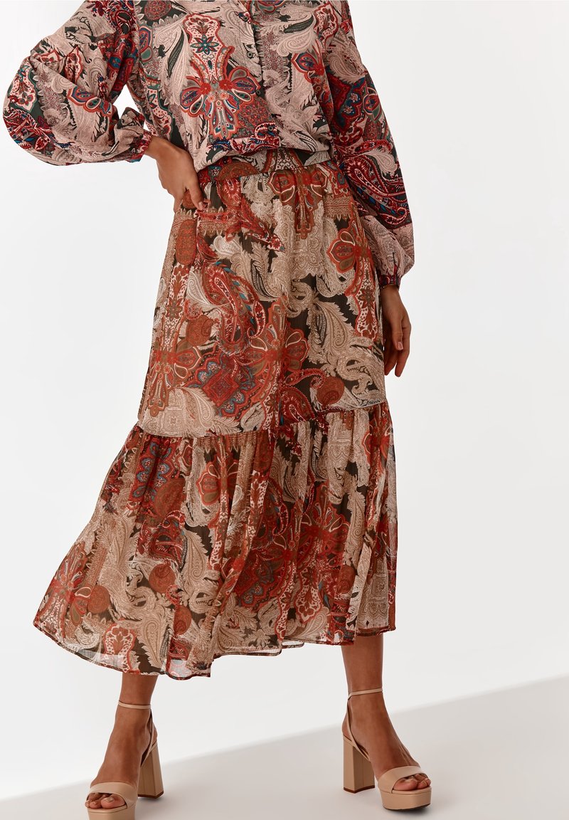 Jupe fluide avec motif cachemire rouge, beige et marron, associée à un chemisier assorti et des sandales beiges à talons hauts sur un mannequin.