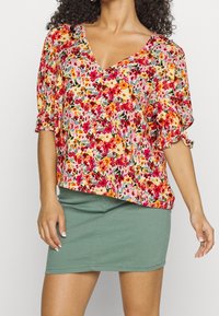 Blouse fleurie à manches courtes bouffantes avec des motifs rouges, orange, jaunes et verts, associée à une mini-jupe verte moulante.