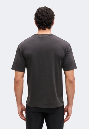 Mand med mørkt, krøllet hår iført en ensfarvet mørkegrå kortærmet t-shirt og sorte bukser, vendt væk mod en ensfarvet baggrund.