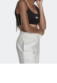 Kvinna i svart kort tanktop med vit logga och högmidjade vita byxor med elastisk midja och snörning, mot ljus bakgrund.