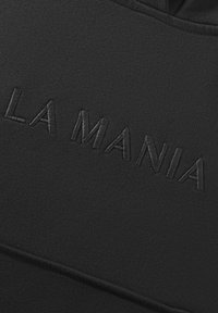 Czarna tkanina z fakturowanym, haftowanym logo "LA MANIA" w lekko wypukłym wzorze, tworzącym subtelny kontrast z materiałem.