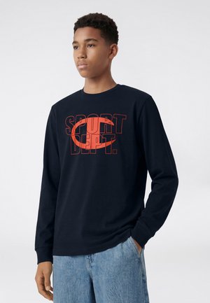 Jeune homme portant un t-shirt noir à manches longues avec texte rouge "SPORT DEPT." et un jean bleu clair, debout avec une main dans la poche.