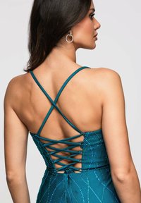 Femme aux longs cheveux foncés porte une robe sarcelle avec des bretelles croisées et lacées dans le dos, ainsi que des boucles d'oreilles pendantes en cristal circulaire.