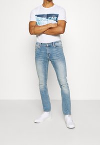 Bílé tričko s modrým grafickým nápisem, světle modré úzké džíny a bílé tenisky. Hladká textura a ležérní design.