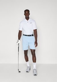Fehér, rövid ujjú polo ing fekete logóval, világos kék rövidnadrág, sötét öv, szürke sportcipő és fehér golfkesztyű, golfütőt tartva.
