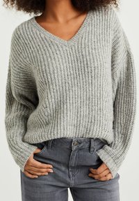 Hellgrauer Strickpullover mit V-Ausschnitt, geprägt von einer gerippten Struktur und weiten Ärmeln, kombiniert mit hoch taillierten grauen Jeans.