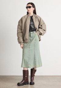 Overdimensjonert khaki bomberjakke, svart grafisk t-skjorte, lysegrønn denimskjørt og brune chunky hæler. Klare solbriller og rett hår.