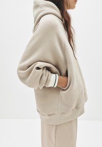 Beige übergroßer Hoodie mit einer Kordelzugkapuze, Fronttasche und gerippten Bündchen mit weißen und grünen Streifen. Weicher, strukturierter Stoff.