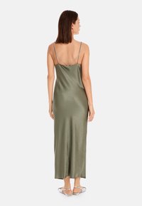 Langes olivgrünes Satin-Kleid mit dünnen Trägern und offenem Rücken, das über eine glatte Textur und einen geraden Saum verfügt, getragen mit silbernen Sandalen.