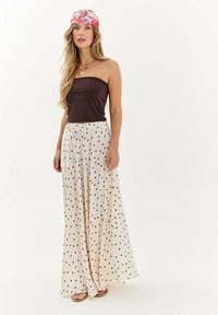 Mladá žena v hnedom topi bez ramienok, smotanovej maxi sukni s bodkami, s ružovou šatkou a sandálmi, stojí pred jednoduchým bielym pozadím.