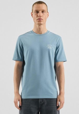 Jonge man met kort haar draagt een lichtblauw T-shirt met kleine witte tekst "Quite Sense" op de linkerborst, gecombineerd met een donkere jeans.