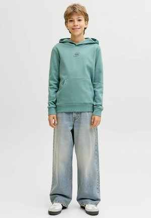 Junge Person steht und trägt einen türkisfarbenen Kapuzenpullover, weite hellblaue Jeans und weiße Sneakers vor einem schlichten hellen Hintergrund.