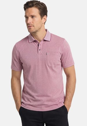 Mężczyzna ubrany w jasnofioletową krótką koszulkę polo z zapięciem na guziki i kieszenią na piersi, ręce w kieszeniach czarnych spodni, patrzący w lewo.