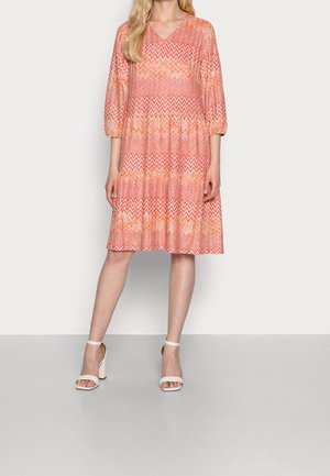 Femme portant une robe mi-longue à motifs zigzag orange et rose, manches trois-quarts, et des sandales à talon bloc blanches avec bride à la cheville.