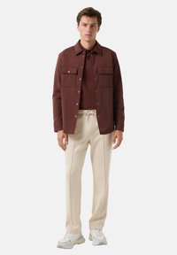 Hnědá košile s knoflíky a kapsami na prsou, nošená přes hnědý top, v kombinaci s béžovými kalhotami a bílými teniskami.