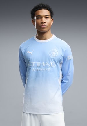 Joven de pie mirando hacia adelante con una camiseta de fútbol de manga larga del Manchester City en azul claro y blanco con logotipos de patrocinadores.